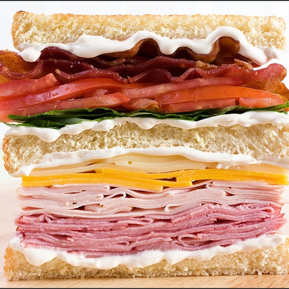 A towering club sandwich with bacon, tomato, lettuce, cheese, ham, and salami on toasted bread.