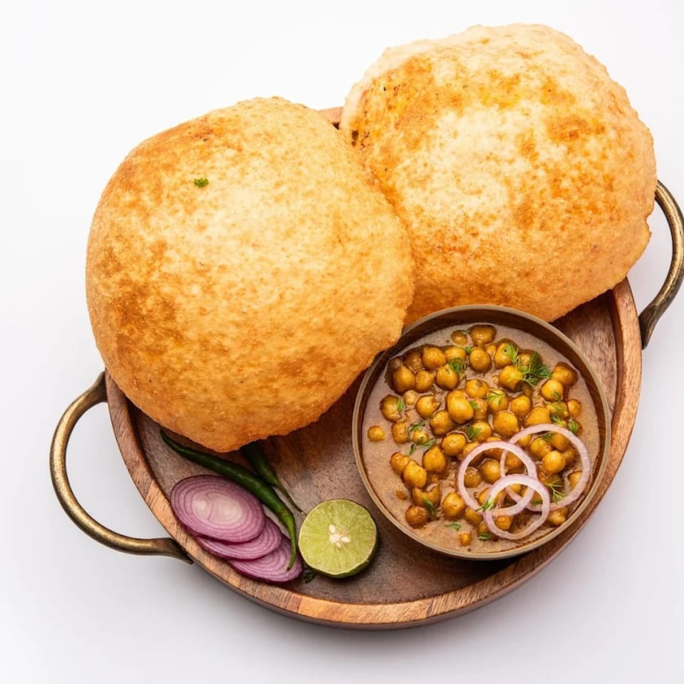 Two golden-brown Indian puri breads with a bowl of chole, red onions, green chilies, and lime.