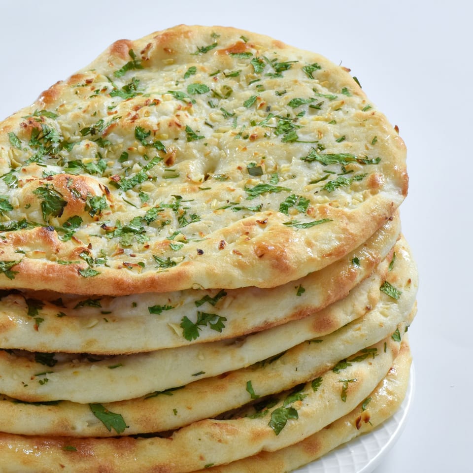 A stack of freshly baked Indian naan bread, garnished with green herbs and garlic.