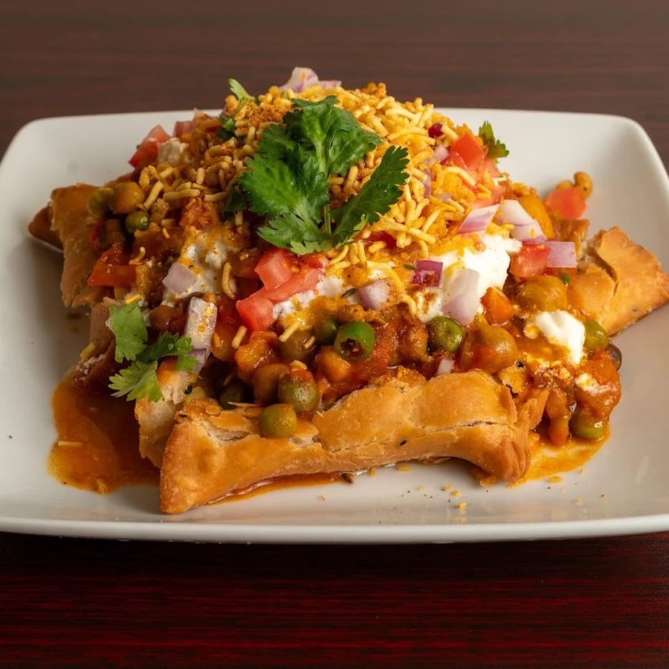 Indian samosa chaat with chickpeas, peas, tomatoes, onions, yogurt, and crispy noodles on a white plate.
