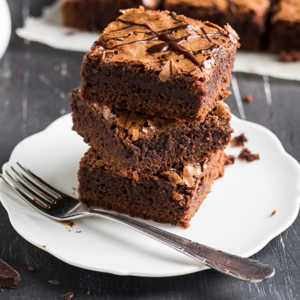 A stack of three rich chocolate brownies with a chocolate drizzle on a white plate with a fork.